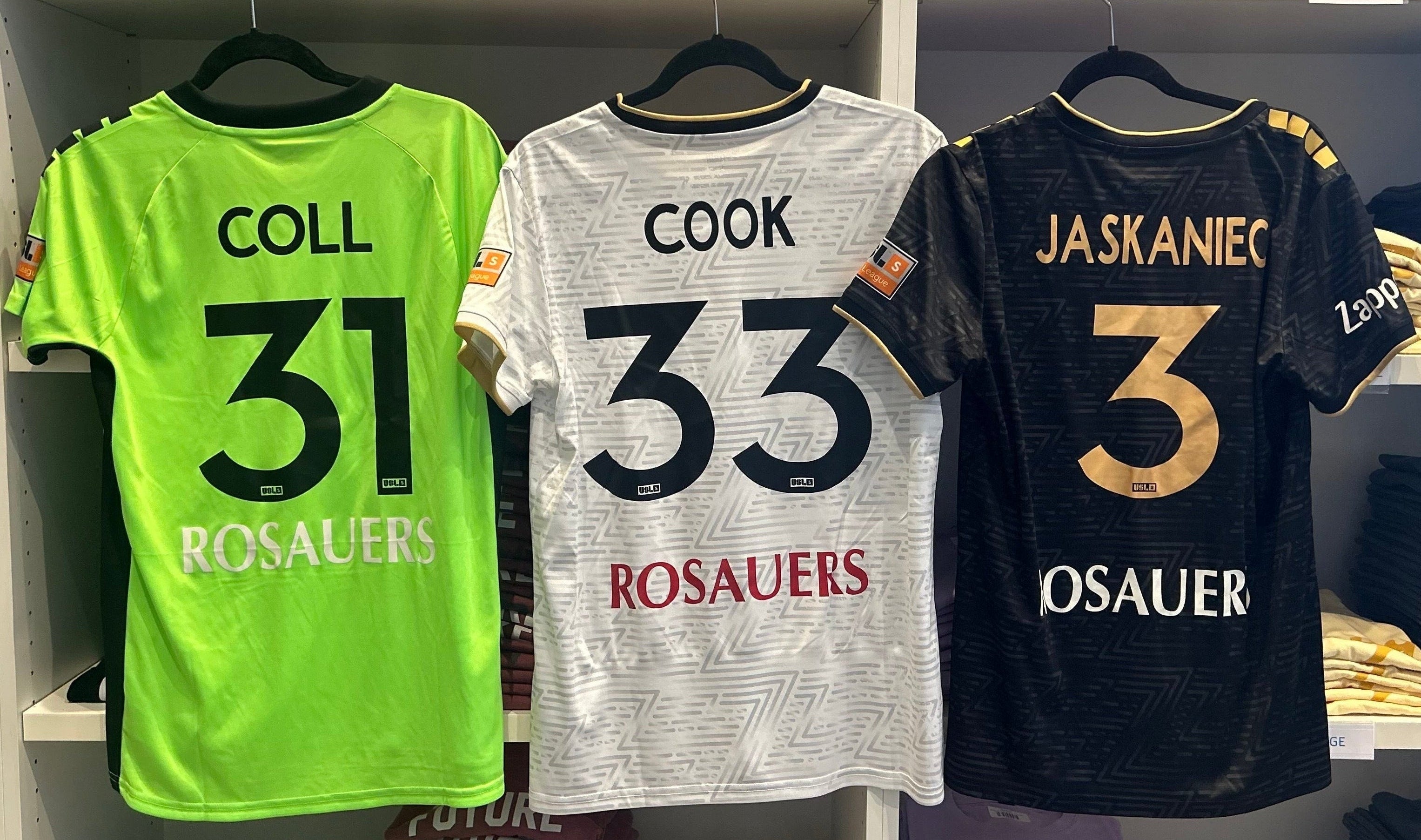 Shelves displaying soccer jerseys with numbers and names in a store setting. From left to right: Coll #31, Cook #33, Jaskaniec #3