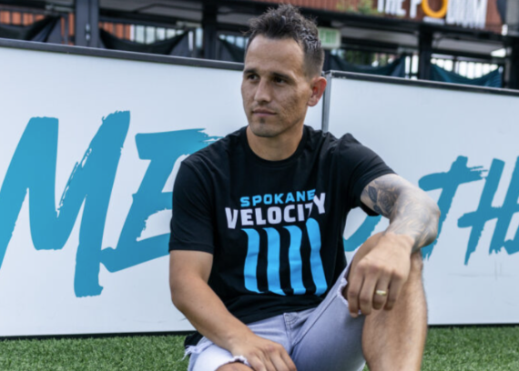 Man sitting on grass wearing a black t-shirt with 'Spokane Velocity' text, white shorts, and white sneakers.