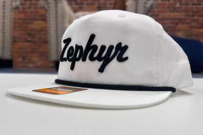 White cap with a black embroidered cursive Zephyr on a white surface with a brick wall background