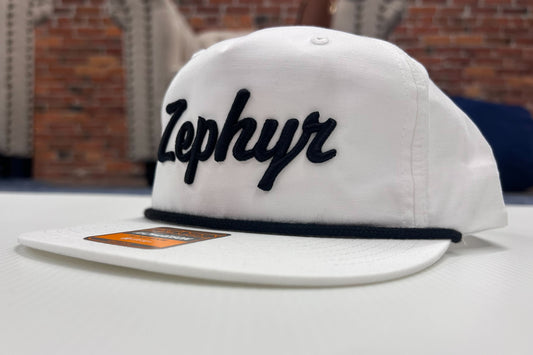 White cap with a black embroidered cursive Zephyr on a white surface with a brick wall background