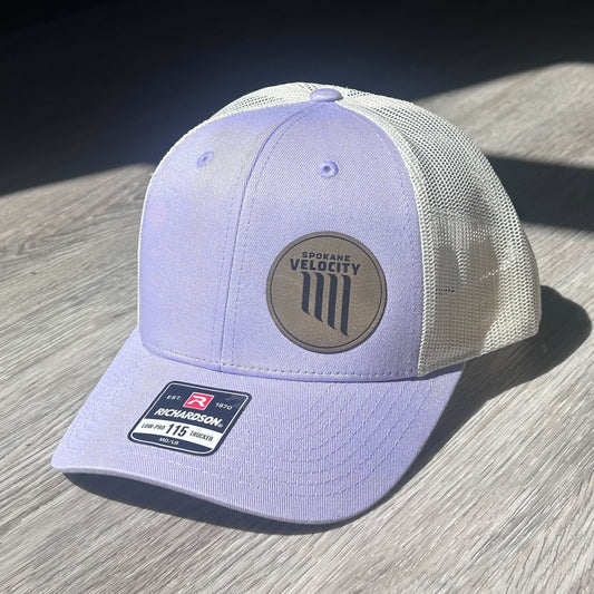 Lilac and cream trucker hat with a Velocity FC logo patch on a wooden surface