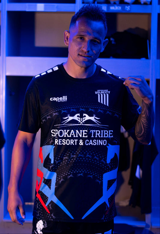 A man wearing a black short-sleeve soccer jersey with a Spokane Velocity crest, 'Spokane Tribe Resort & Casino' and 'Capelli' branding, standing in a locker room under blue lighting.