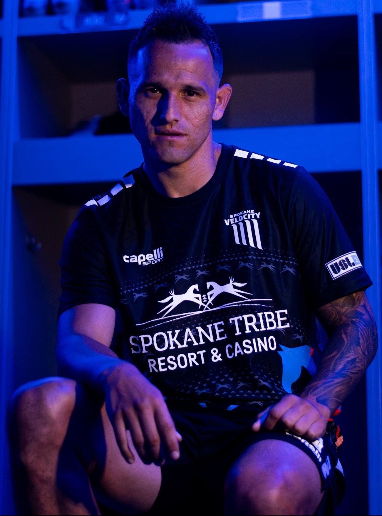 A man wearing a black Spokane Tribe Resort & Casino athletic jersey and shorts, sitting in front of blue-lit lockers.
