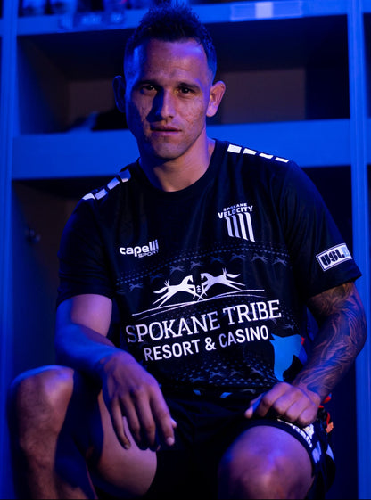 A man wearing a black Spokane Tribe Resort & Casino athletic jersey and shorts, sitting in front of blue-lit lockers.